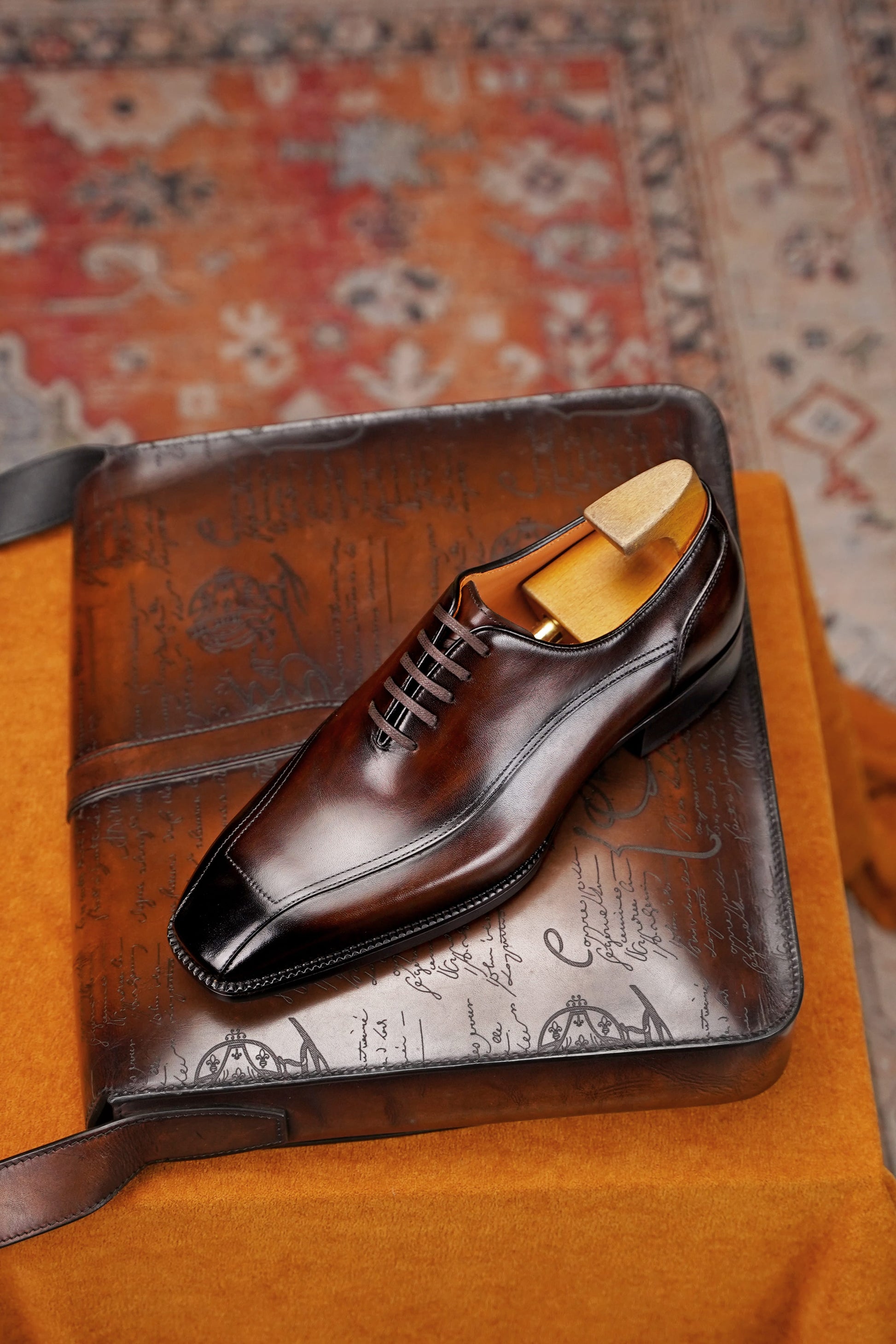 A polished dark brown leather dress shoe with a black toe cap and lace-up front, displayed on a brown leather case over an orange fabric surface with a patterned rug in the background.