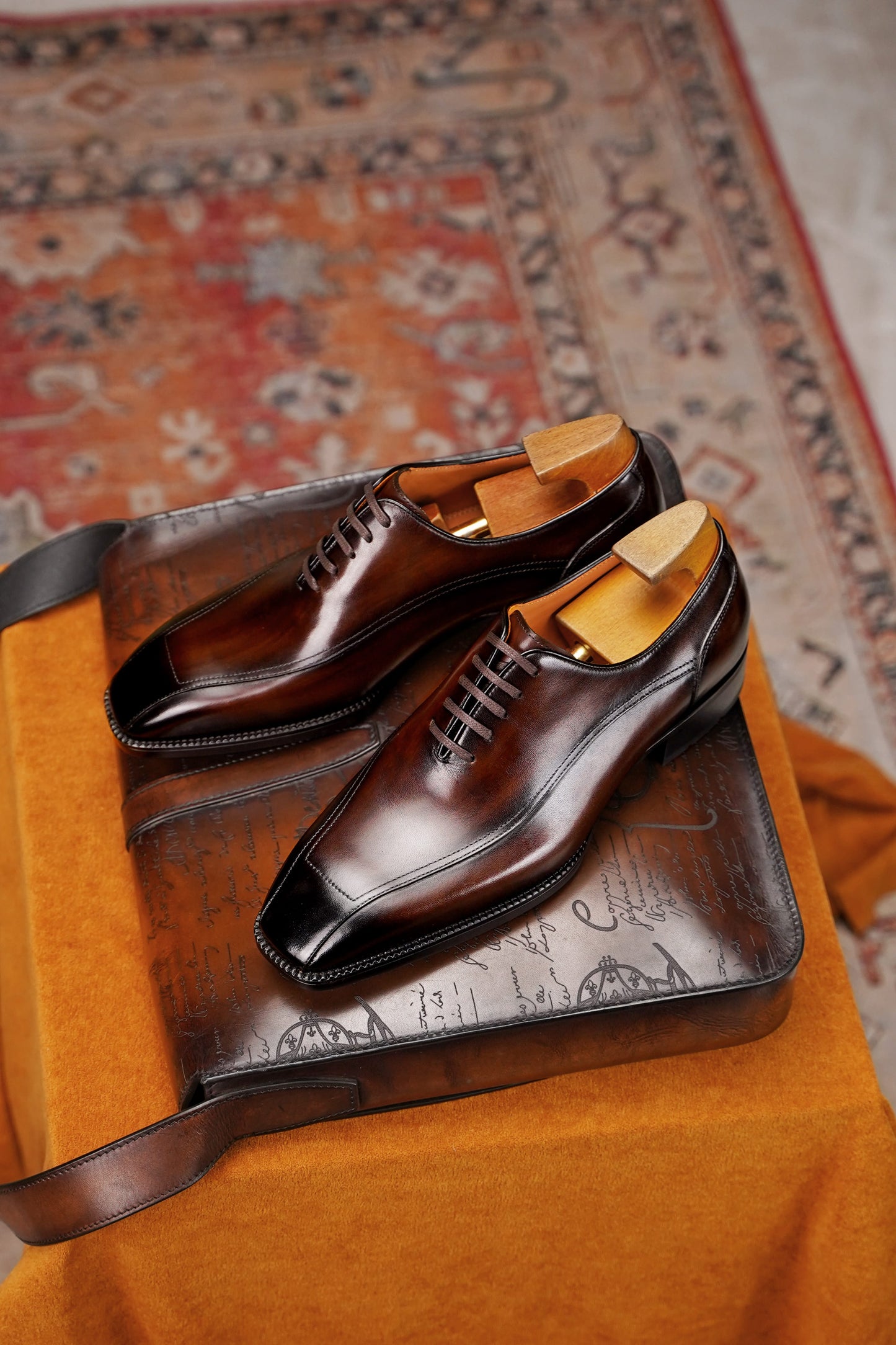 A pair of dark brown leather dress shoes with black toe caps, displayed on a brown leather case atop an orange fabric surface with a patterned rug in the background.