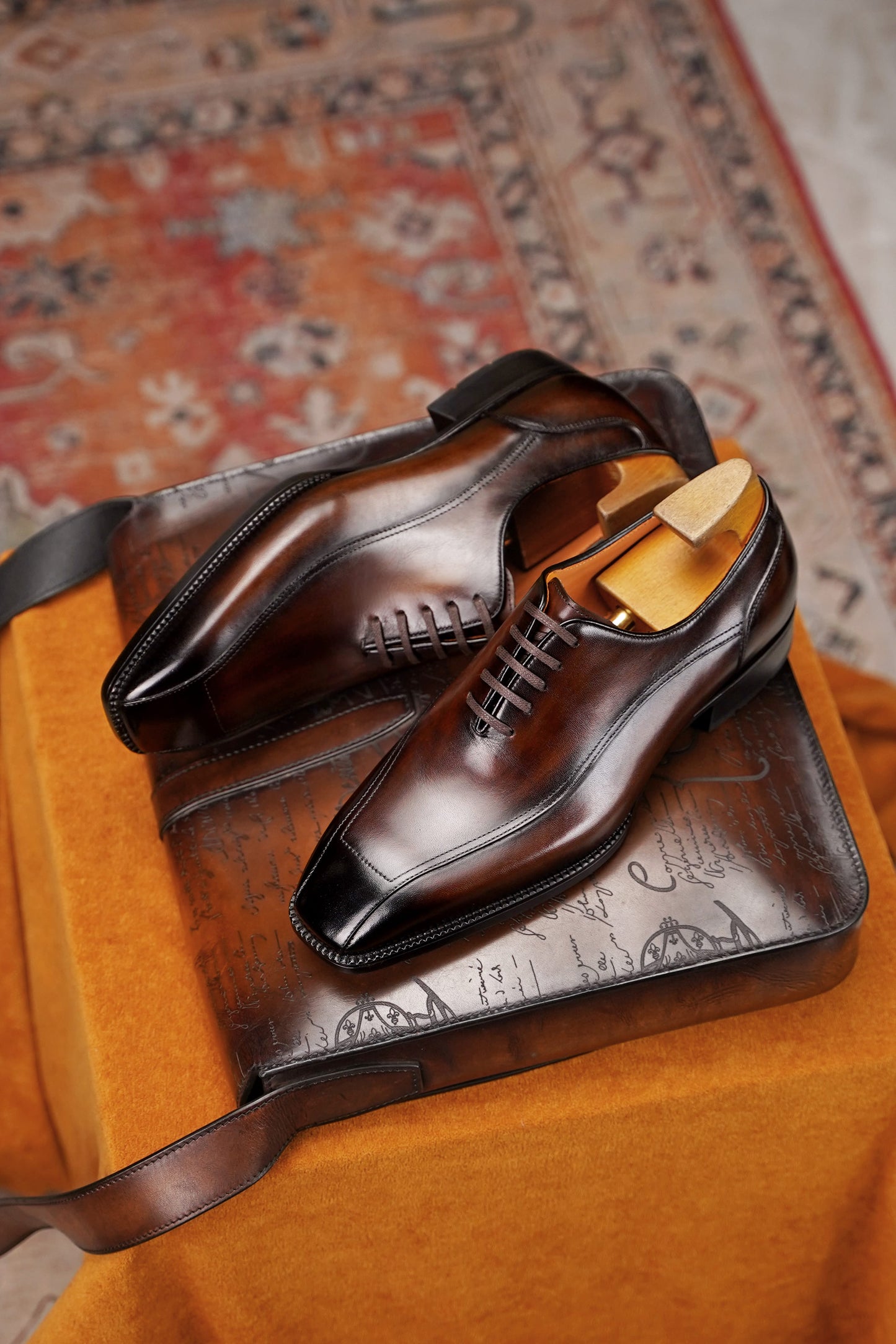 A pair of polished dark brown leather dress shoes with black toe caps and black laces, displayed on a brown leather case over an orange fabric surface with a patterned rug in the background.