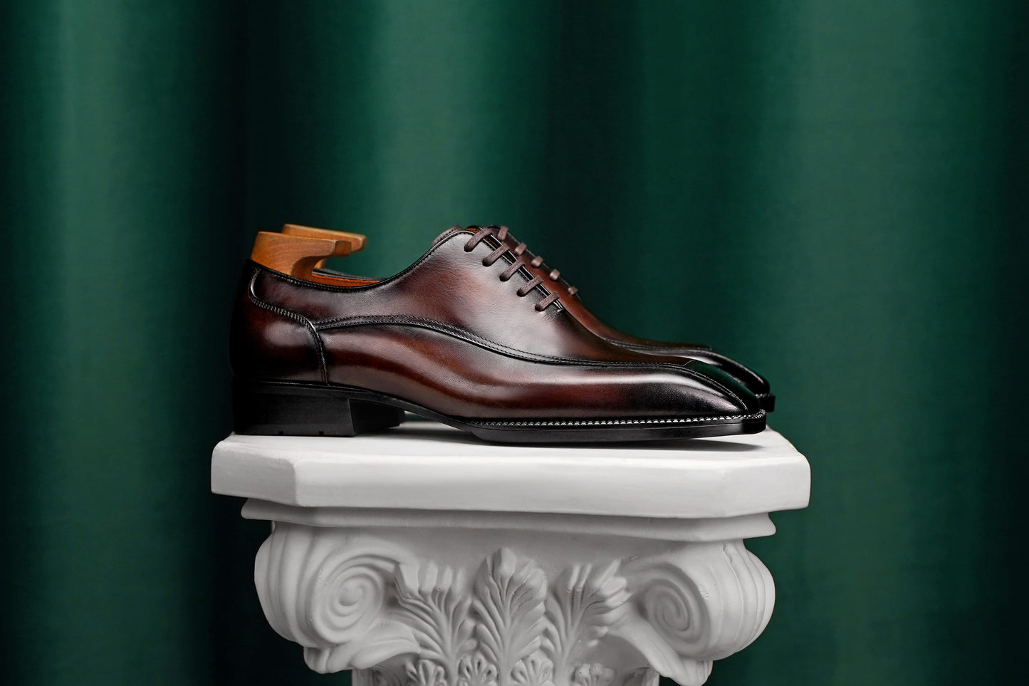 A polished dark brown leather lace-up dress shoe with a glossy finish and black sole, displayed on a white decorative pedestal against a dark green curtain background.