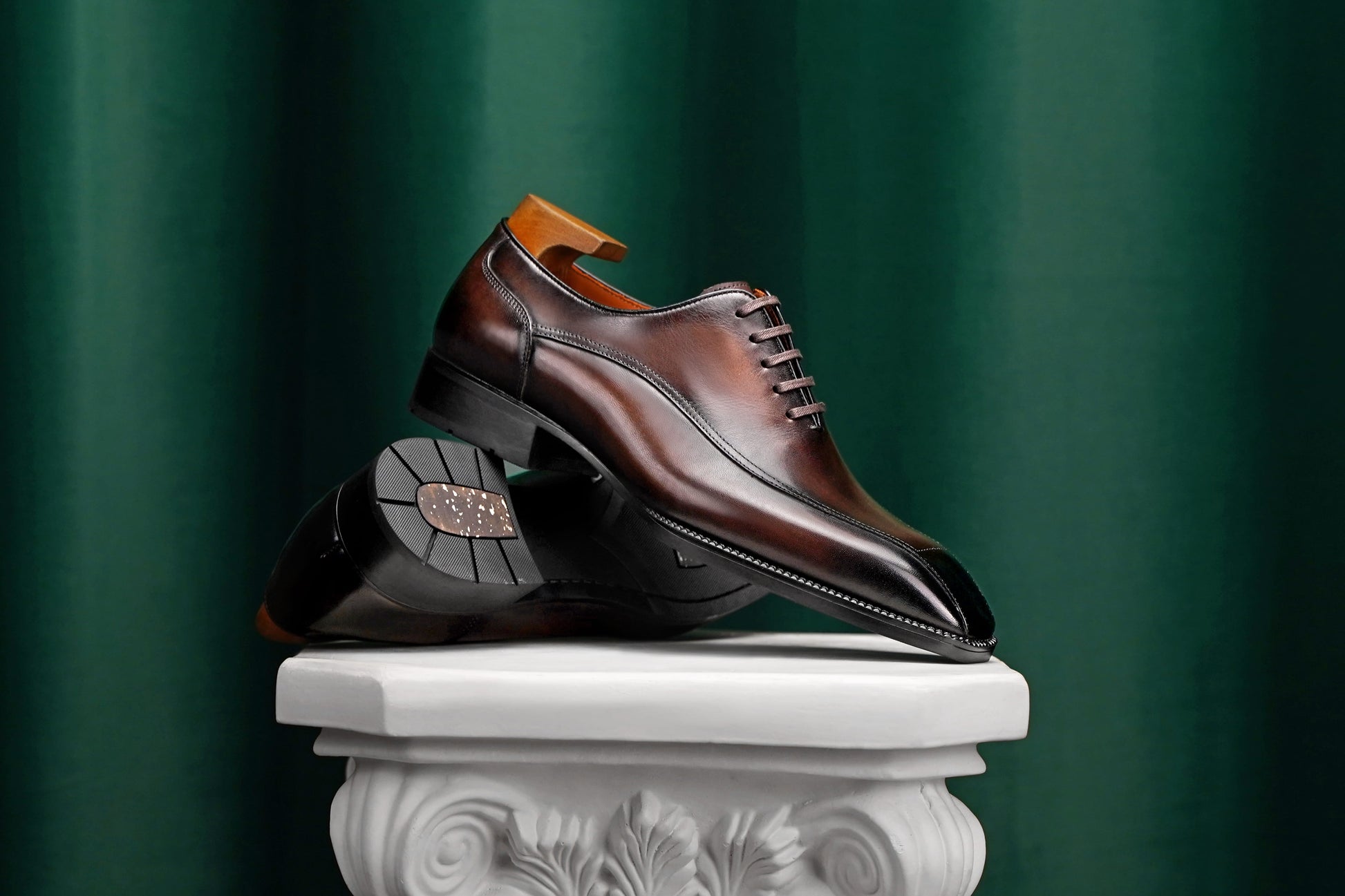A pair of dark brown leather oxford dress shoes with black soles, displayed on a white classical-style pedestal against a dark green velvet curtain background.