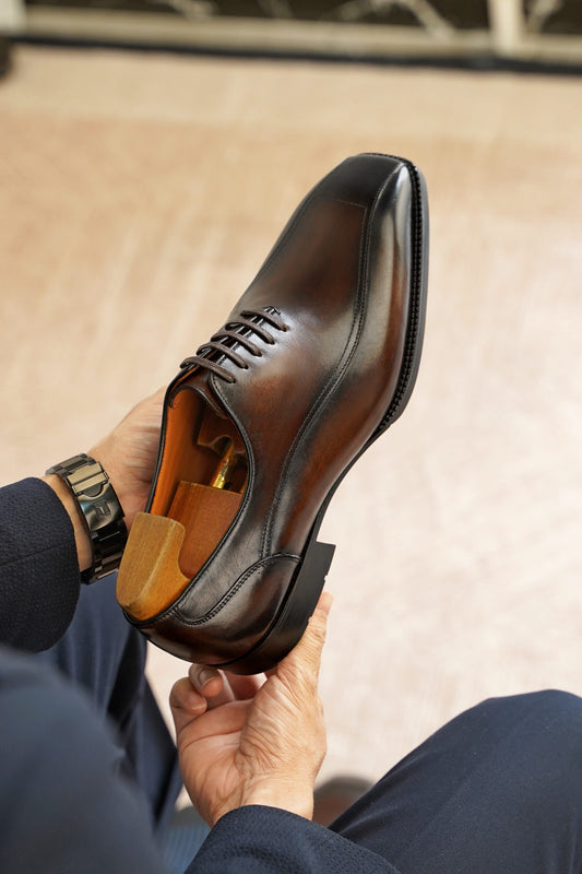 A close-up of a person holding a dark brown leather dress shoe with a wooden shoe tree inside, showing the tan interior lining and laces. The person is wearing navy blue trousers and a gold-toned watch.