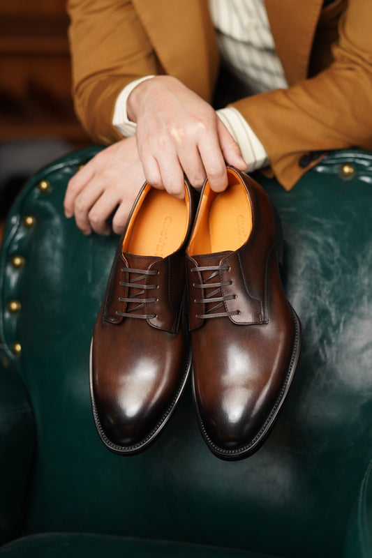 A pair of polished dark brown oxford shoes with lace-up fronts and orange insoles, held by a person wearing a tan blazer and white shirt, seated on a dark green leather armchair.