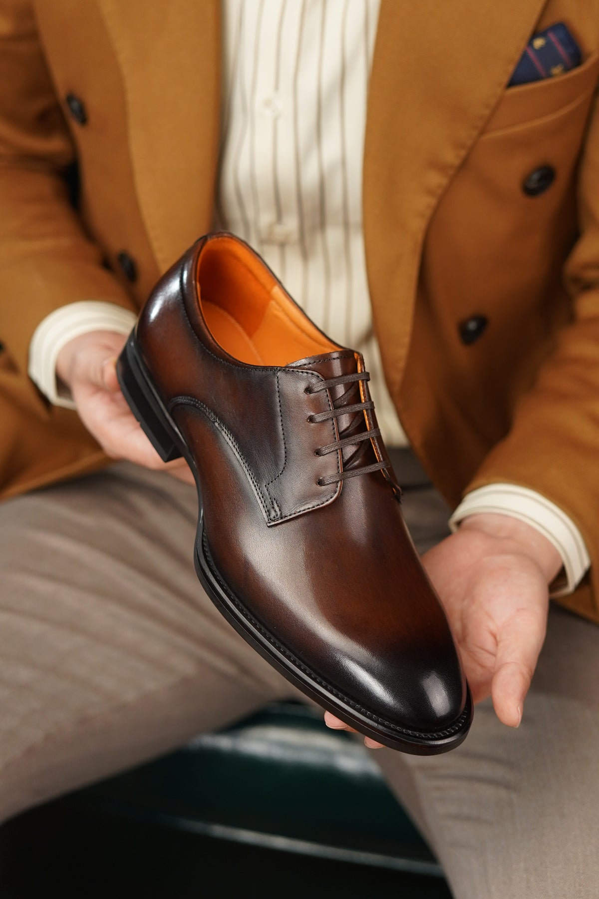 Brown leather dress shoe held by a person wearing a brown coat and beige pants.