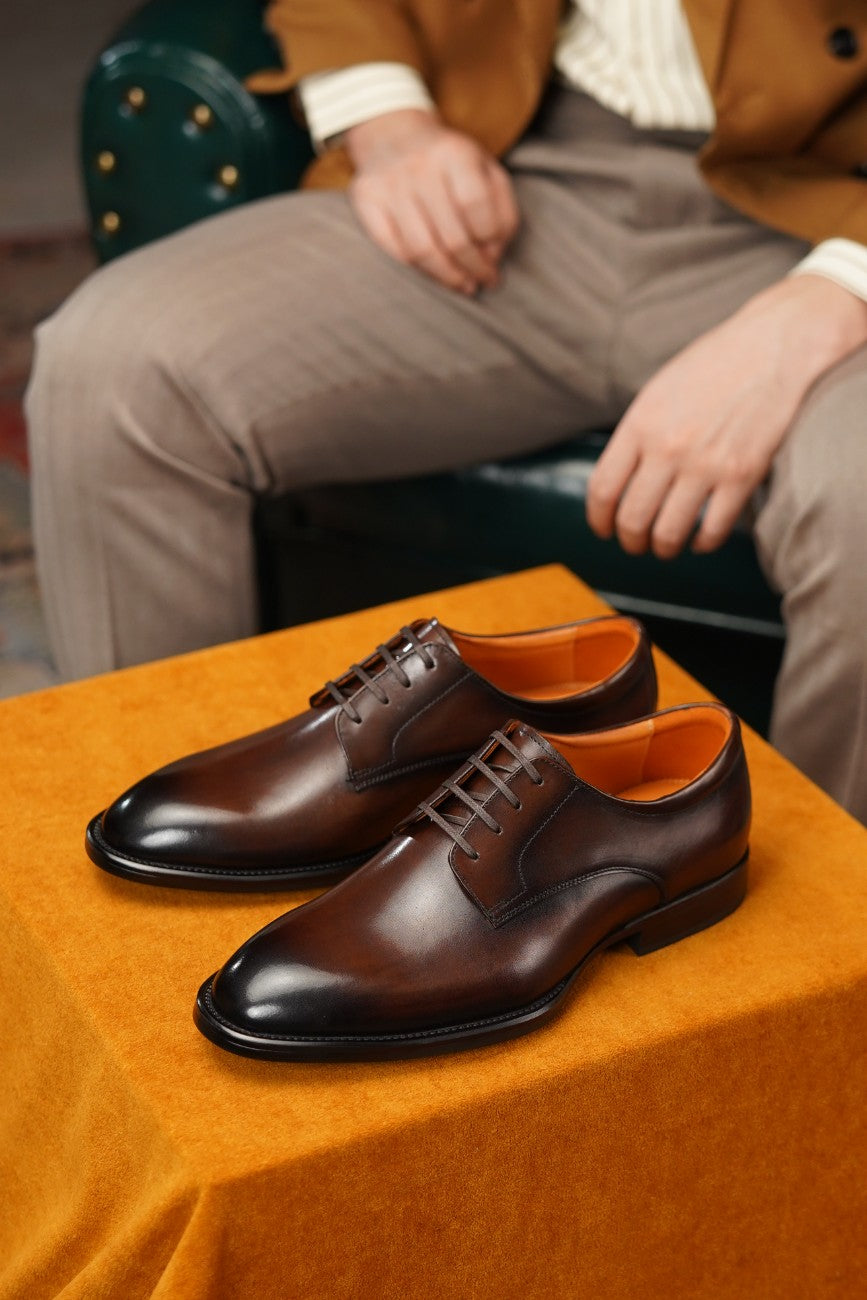 A pair of dark brown leather oxford dress shoes with a glossy finish and black laces, displayed on an orange velvet-covered surface with a model's legs visible in the background.