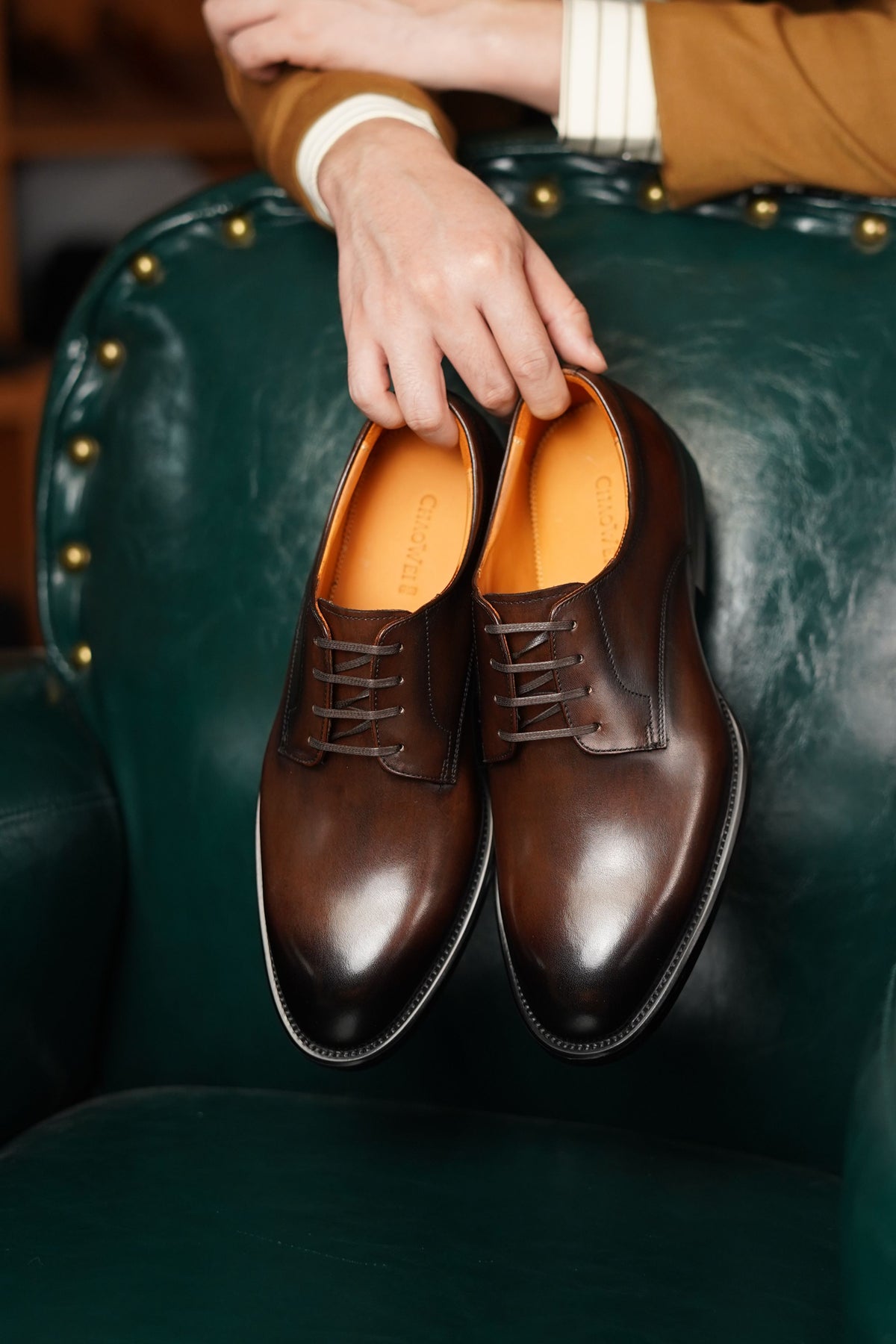 A pair of polished dark brown oxford shoes held by a person's hand against a dark green leather armchair.