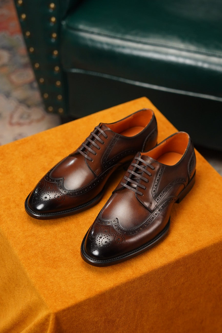 A pair of dark brown leather wingtip brogue dress shoes with a glossy finish, displayed on a textured orange velvet surface with a dark green leather chair in the background.