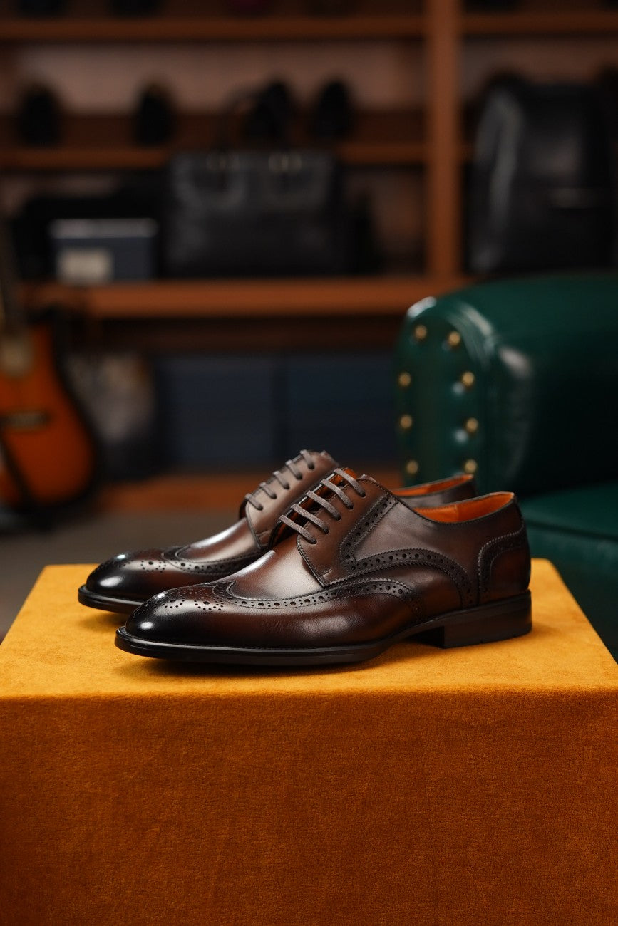 A pair of dark brown leather wingtip derby shoes with decorative brogue detailing, displayed on a textured orange block in a softly blurred boutique setting.