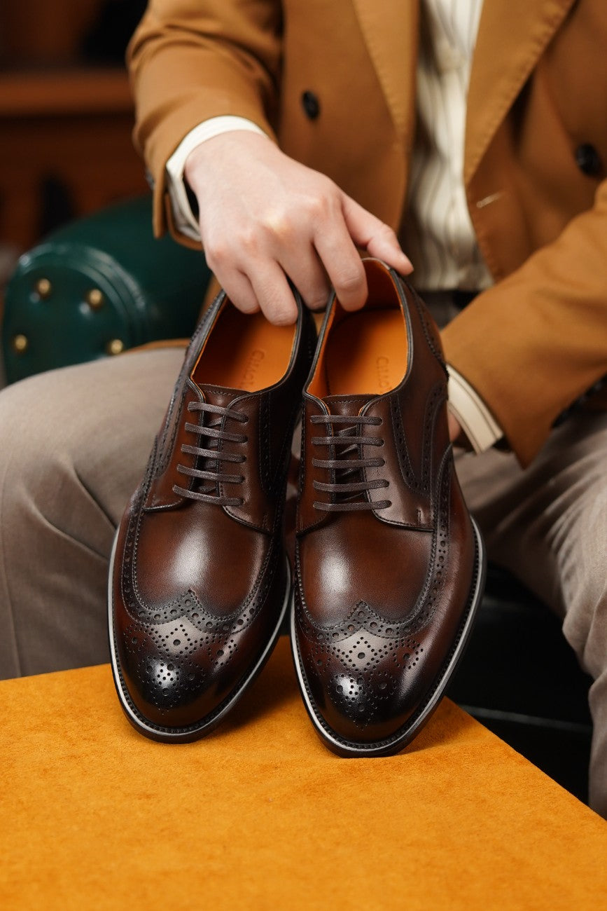 A pair of dark brown leather wingtip derby shoes with brogue detailing, held by a person wearing a tan blazer and beige trousers.