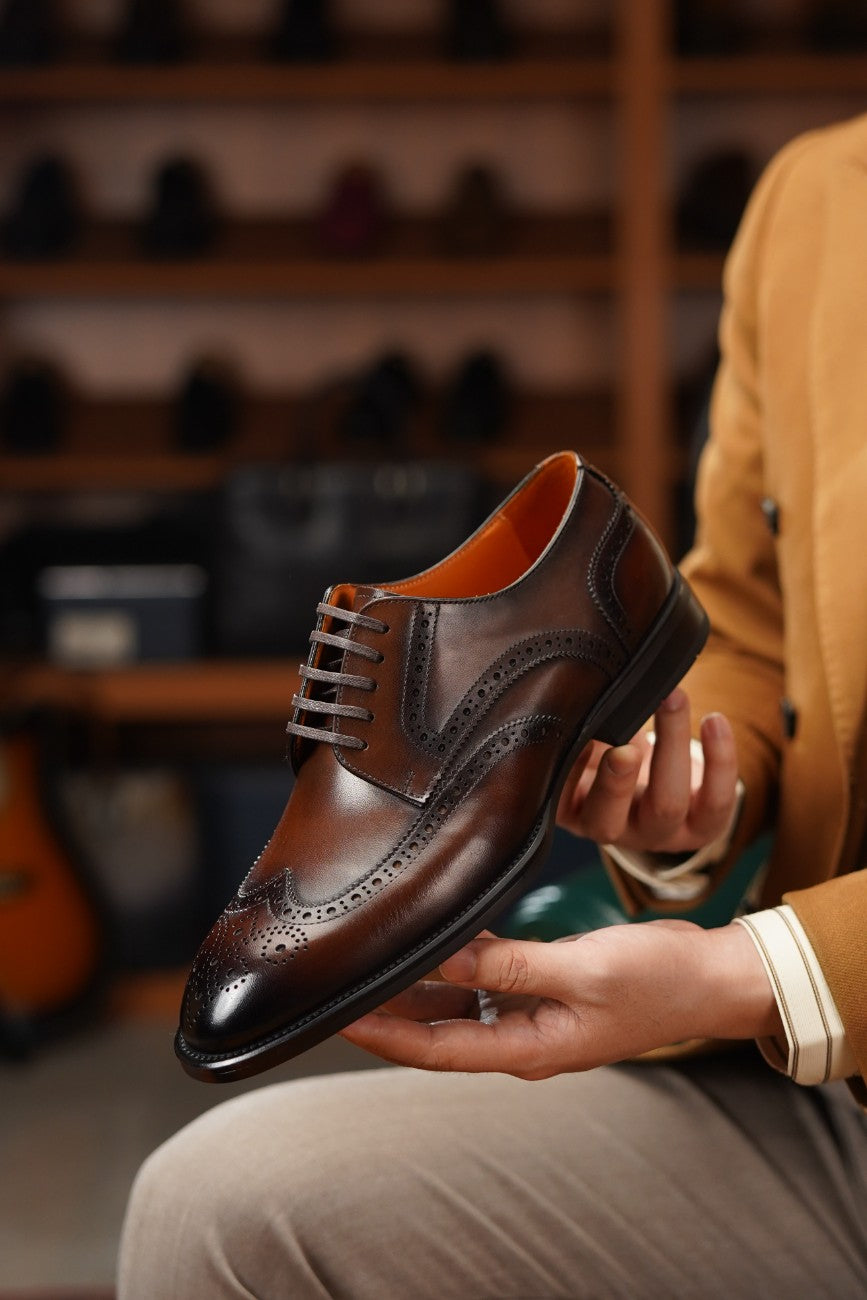 A person in a tan blazer and beige trousers holds up a dark brown leather wingtip derby shoe with brogue detailing and a contrasting orange-brown lining, set against a blurred background of wooden shelves filled with shoes.
