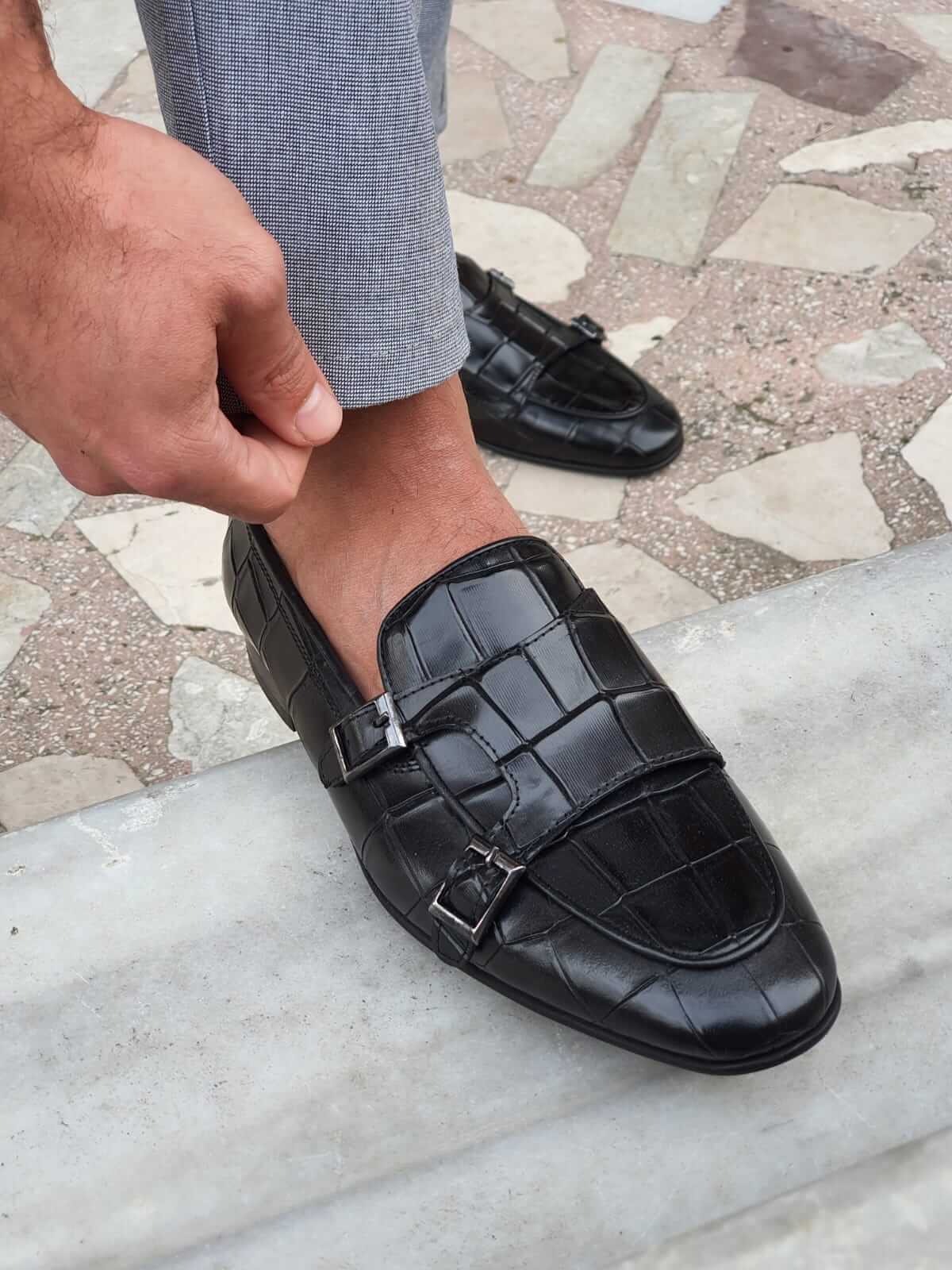 Crocodile-Embossed Double Monk Strap Shoe – close-up of a man adjusting his light grey trousers, highlighting the striking textured leather and sleek buckles. 