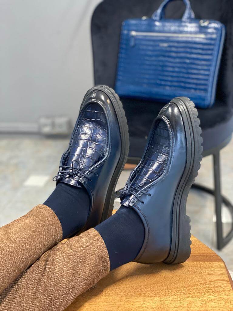 Dark Blue Crocodile Leather Derby Shoes – worn by a man in brown trousers, resting feet on a wooden stool, with a matching blue bag in background, showcasing lasting style.
